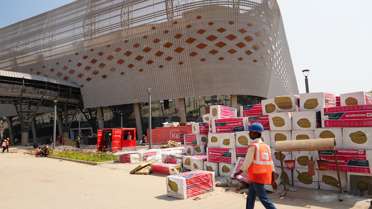 一名穿著安全背心、戴著安全帽的人走過一棟大型金屬建築附近的成堆建築材料。