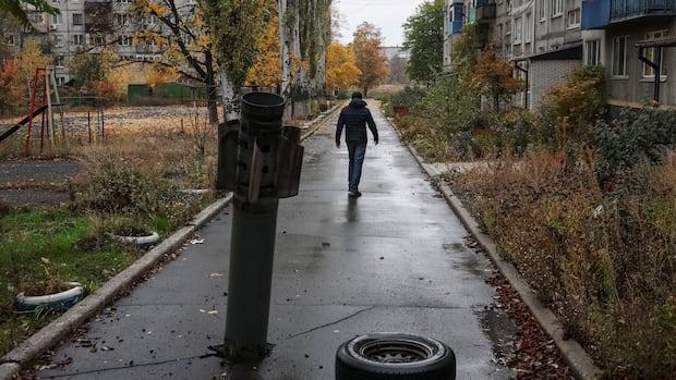 A man is seen walking along a paved path, with a missile embedded in it. Damaged apartment buildings are seen on either side.