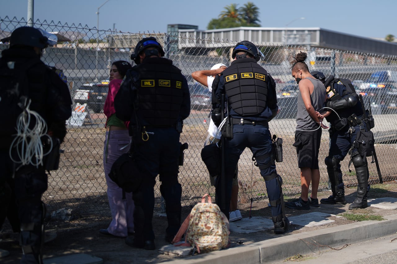 Four officers detain protestors by a wire fence.