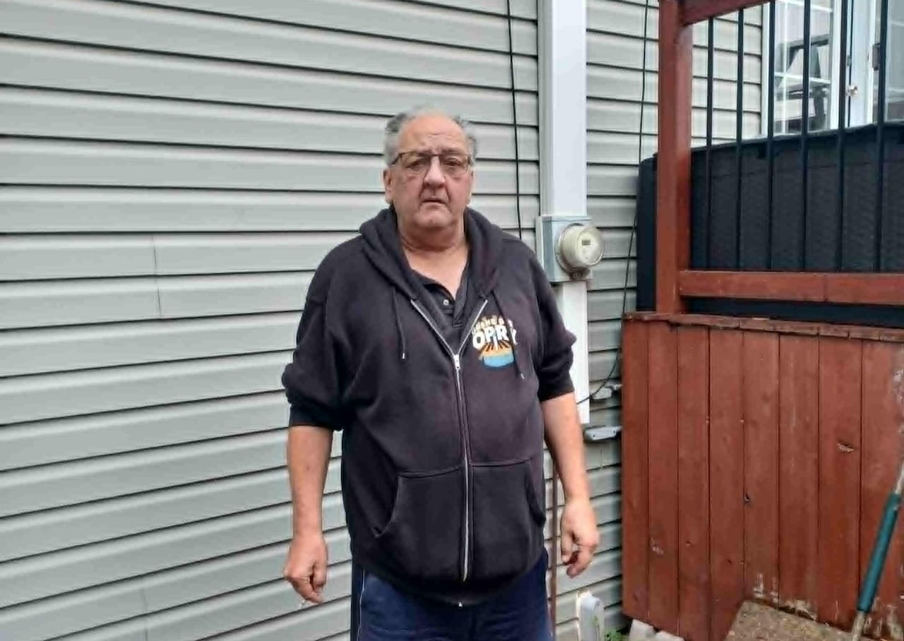 A man stands in a hoodie with a power meter on his house behind him.