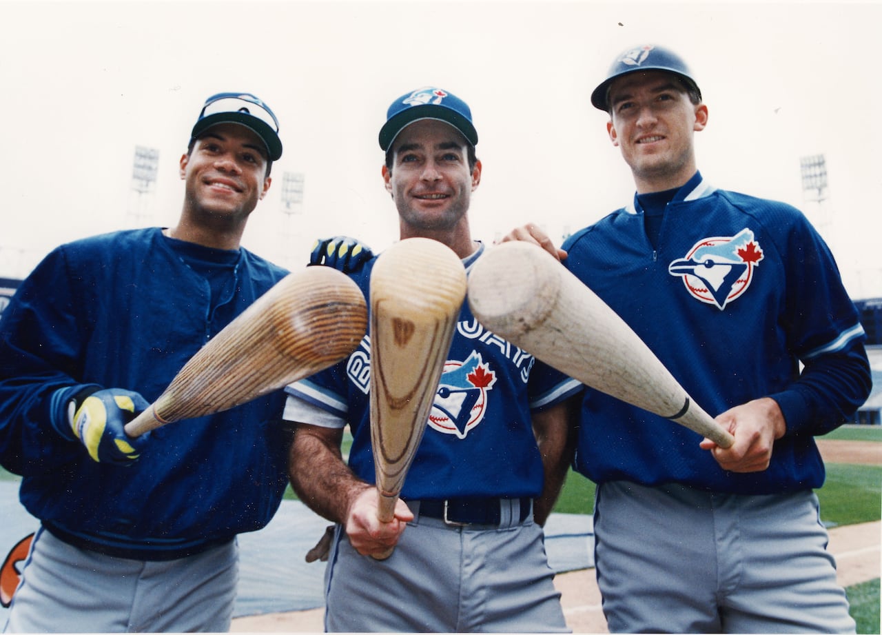 Three baseball players stand alongside one another, each pointing a baseball bat toward the viewer