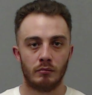 A mug shot of a criminal from dawson creek, he is a man in a white tshirt and tired eyes with a beard