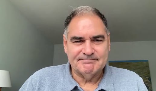 A man smiles while sitting in a room, with a lamp visible.