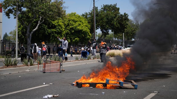 Demonstrators shout and film at police during a protest in the Paramount section of Los Angeles, Saturday, June 7, 2025, after federal immigration authorities conducted operations. (AP Photo/Eric Thayer) Protesters shout and film at police on a street as debris burns in the foreground.