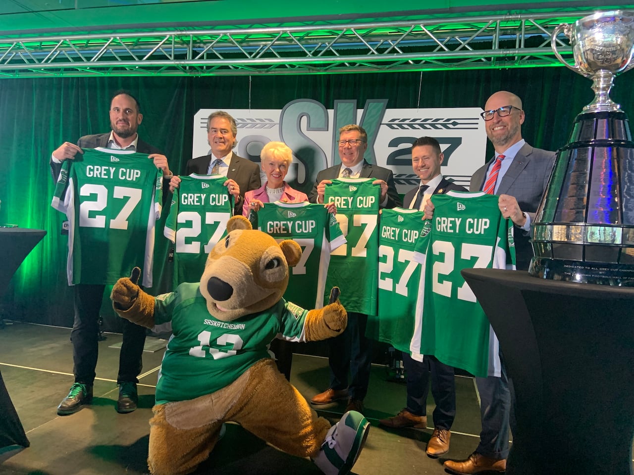 Six people hold up green and white football jerseys with the number 27 on them. A mascot kneels in front