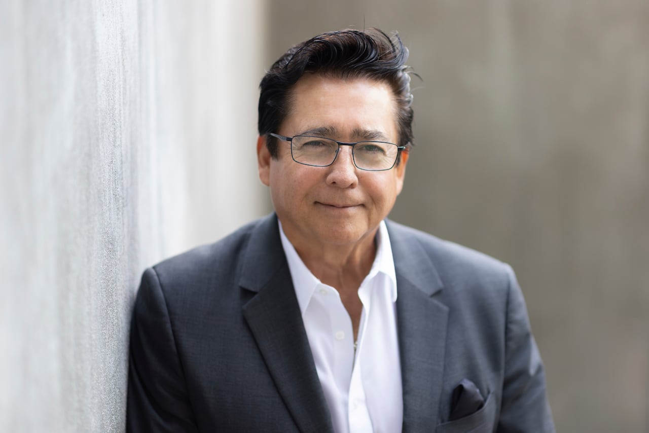 A man in a suit jacket with glasses and black hair smiles at the camera.