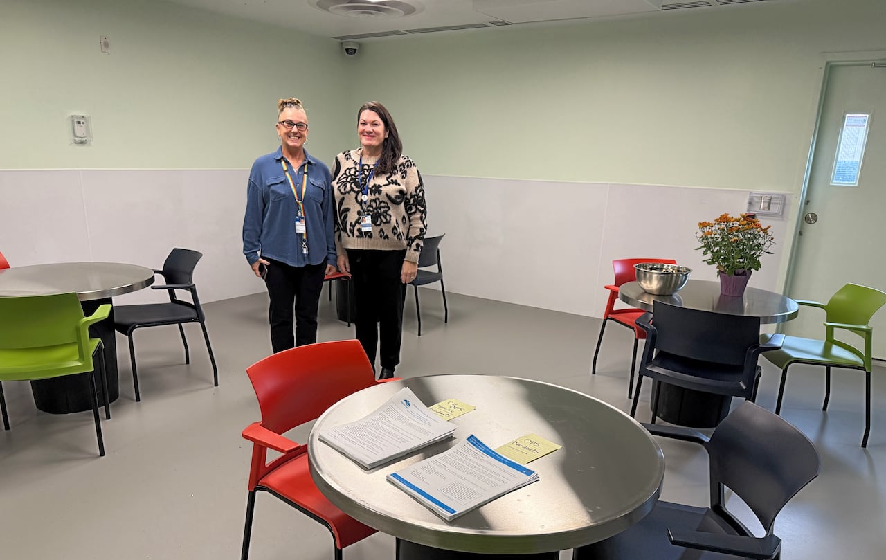Two women pose for a photo, standing inside a room with round metal tables and colourful plastic chairs. 