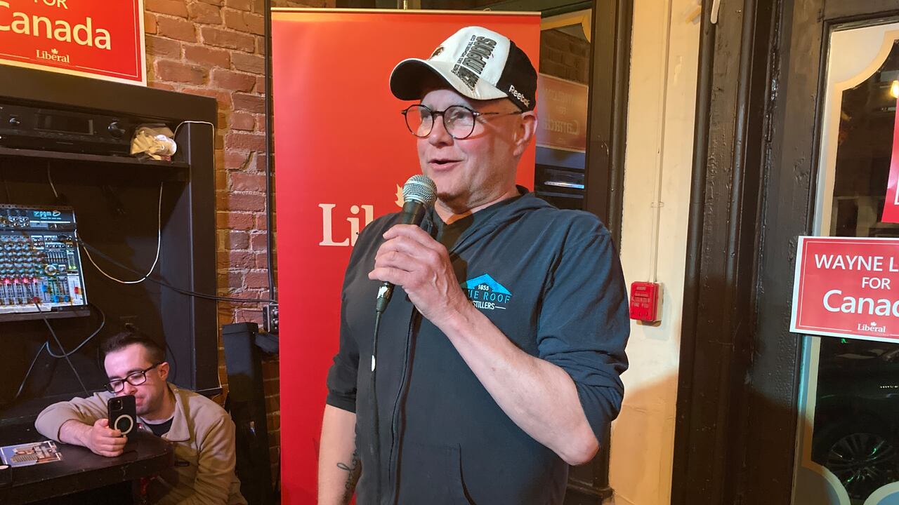 A antheral   successful  a achromatic  sweater and ballcap speaks into a microphone successful  beforehand   of a reddish  run  sign.