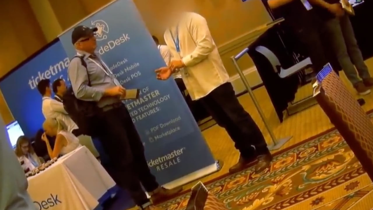 An undercover journalist talks to a man with a blurred face on the floor of a ticketing industry convention.