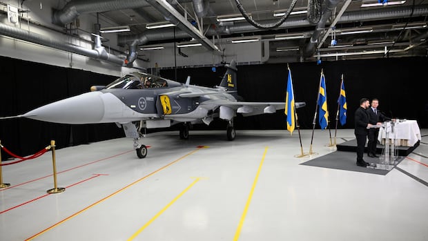 A fighter jet is pictured in an industrial space. Two men stand in front of it behind a podium.