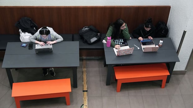 Three young people sit at tables, staring at laptops.