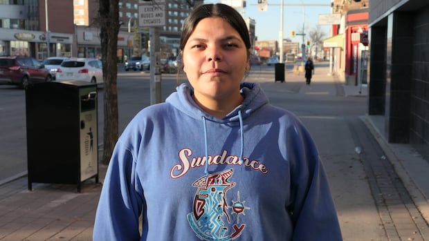 A young woman in a blue hooded sweatshirt with dark hair pulled back. She stands outside on a city sidewalk 
