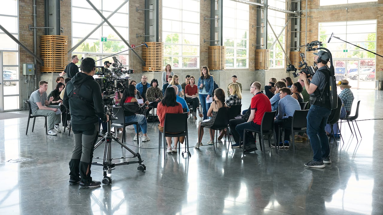 A wise shot of the set which shows the guest, the interviewers and the production team around them. All are in a bright studio with natural light.