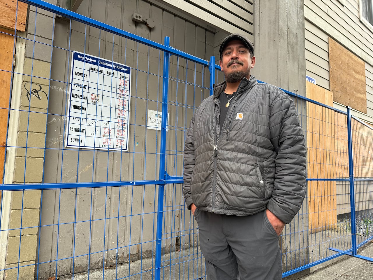 A man with a goatee and a cap stands in front of a boarded-up building.