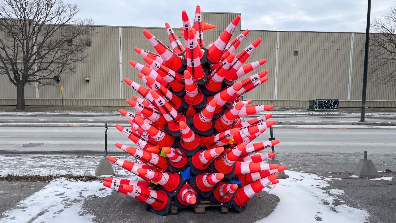 Is this the most Montreal Christmas tree ever?; A christmas tree made entirely of orage road safety cones sits in a parking lot by a street in an industrial or commercial area