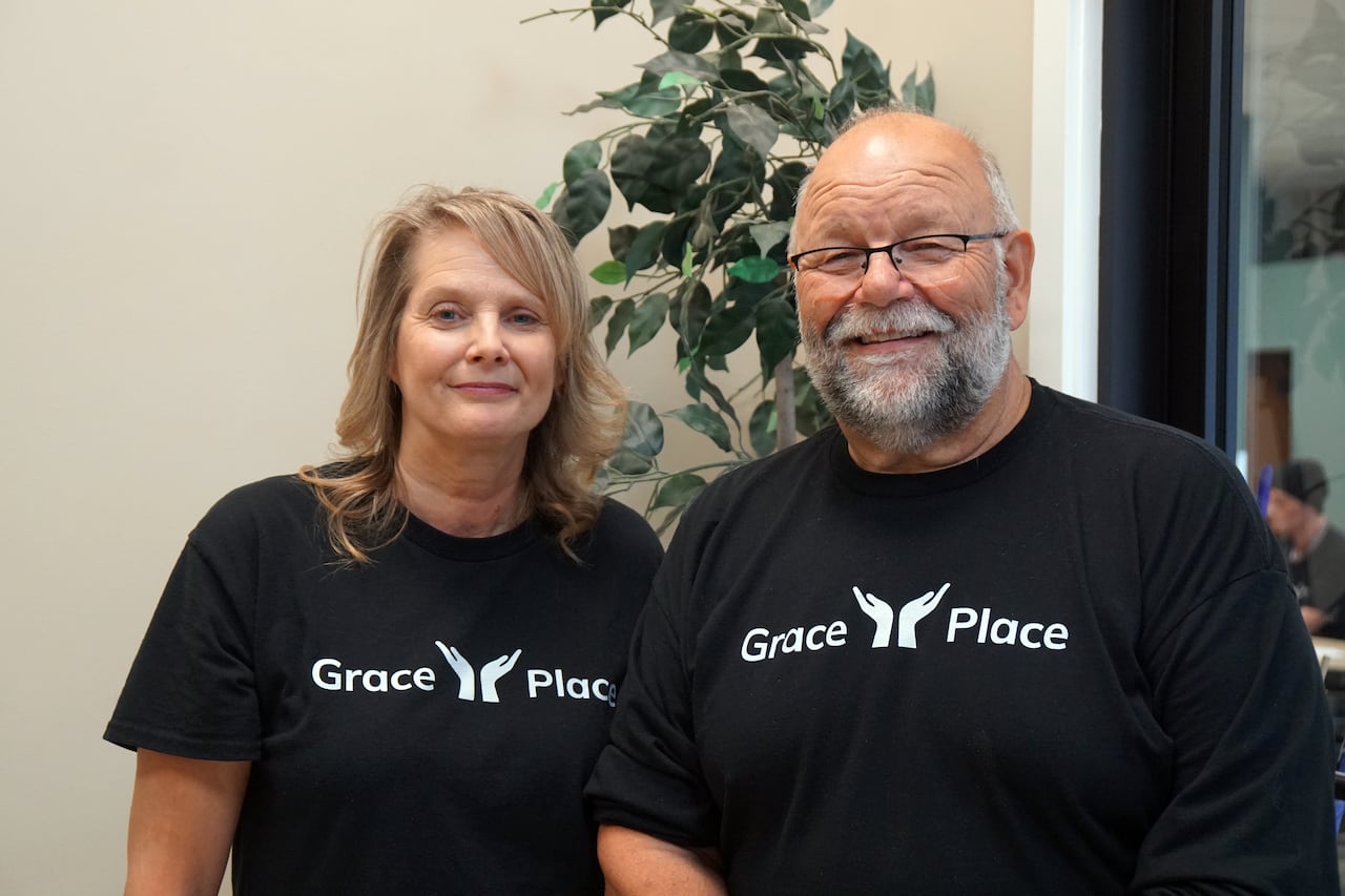 Two people wearing black t-shirts which say 'Grace Place' on them in white are seen standing beside each other.