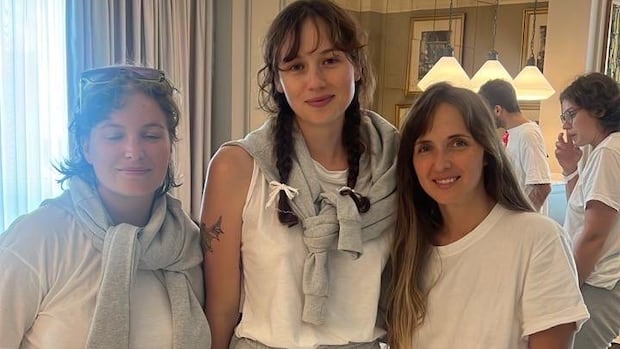Three smiling women stand in a hotel lobby. They're all wearing white t-shorts, grey sweatpants and sandals.