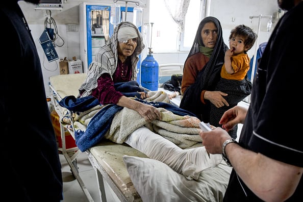 An older woman sits on a stretcher in a hospital. A bandage covers her head and eye. A woman holding a toddler looks on.