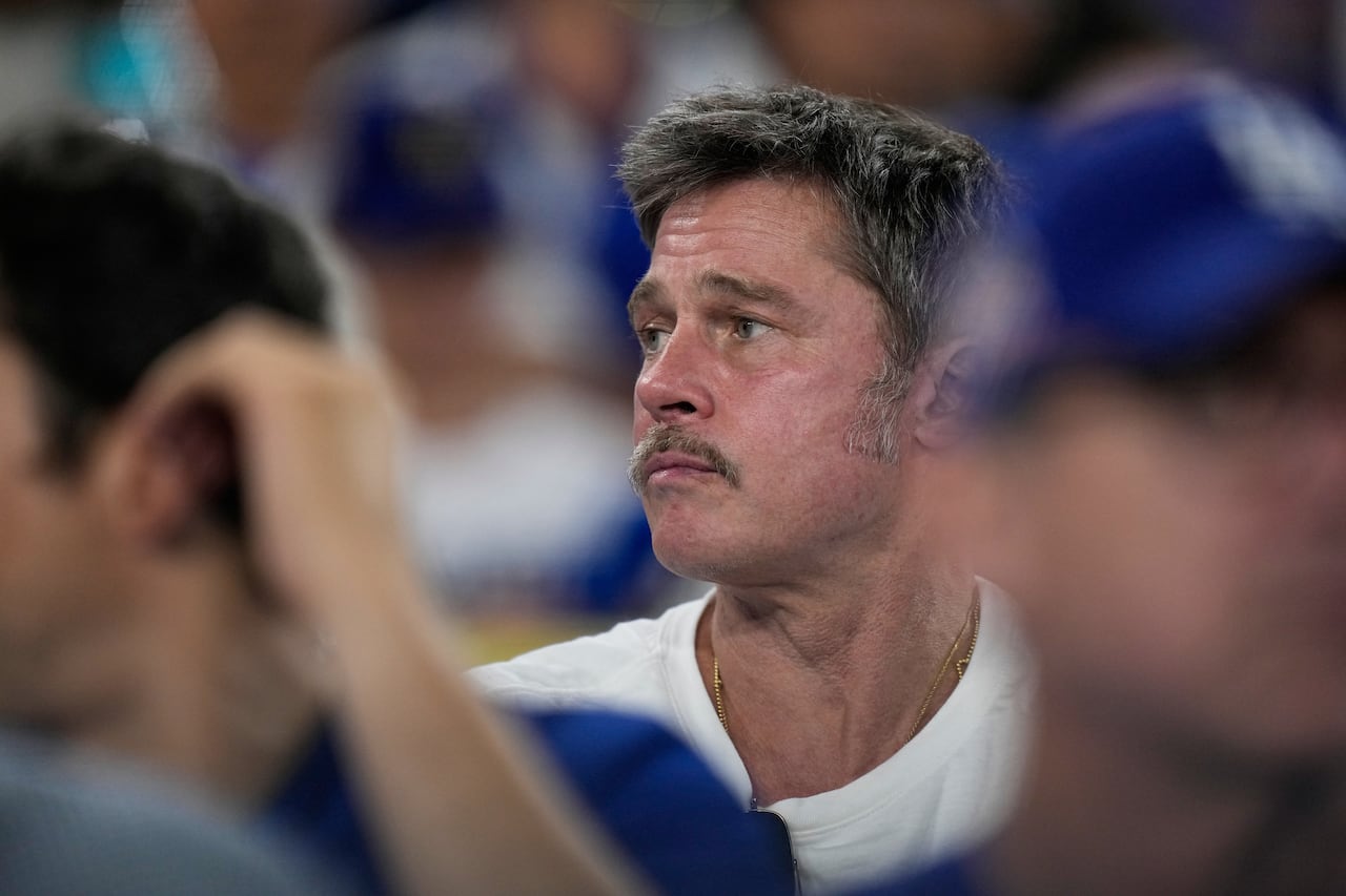 A man with a mustache, wearing a white t-shirt, is seen sitting in the stands of a stadium.