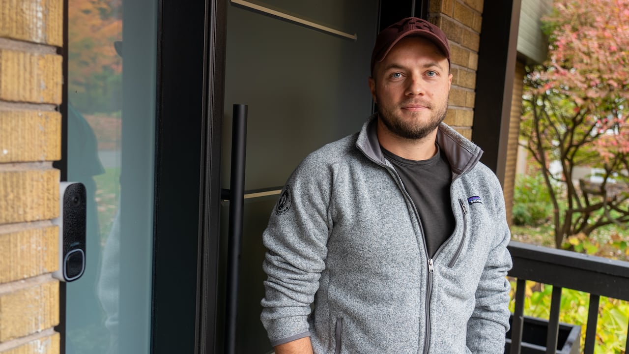 A man in a gray zip-up sweater stands on the porch.