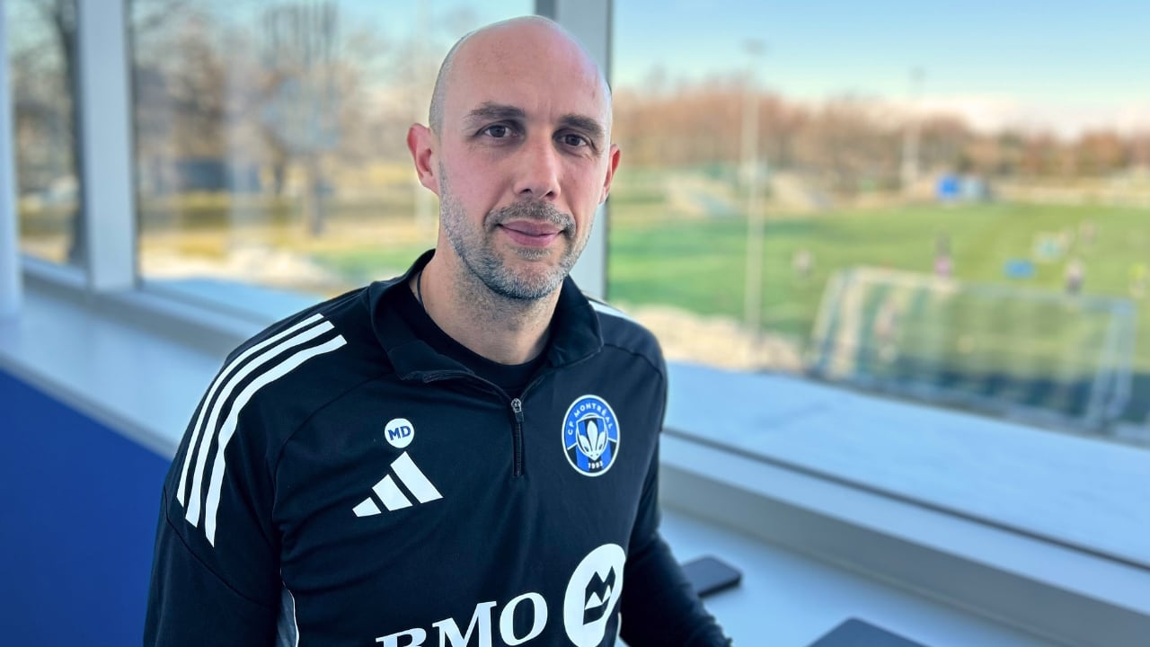 Marco Donadel poses for the camera in a tracksuit in front of a window with a soccer field outside