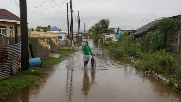 A man wades through a flooded street ahead of the forecasted arrival of Hurricane Melissa in Old Harbour, Jamaica, Monday, Oct. 27, 2025. (AP Photo/Matias Delacroix) A man wearing a hooded sweatshirt and shorts stands while holding a bicycle while in an alley filled with water up to his ankles.