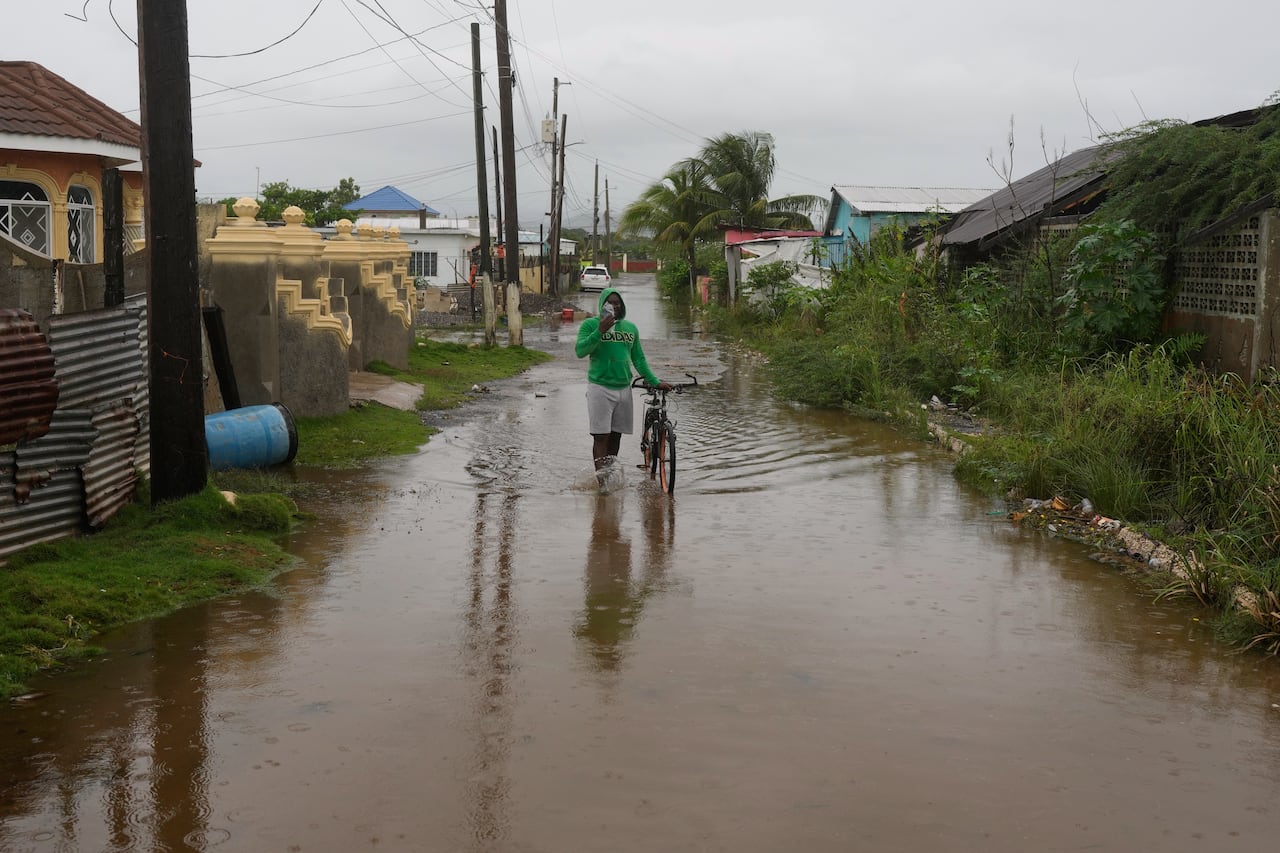 Jamaica, Cuba Brace for Hurricane Melissa to Make Landfall Today 2 A man wearing a hooded sweatshirt and shorts stands while holding a bicycle while in an alley filled with water up to his ankles.
