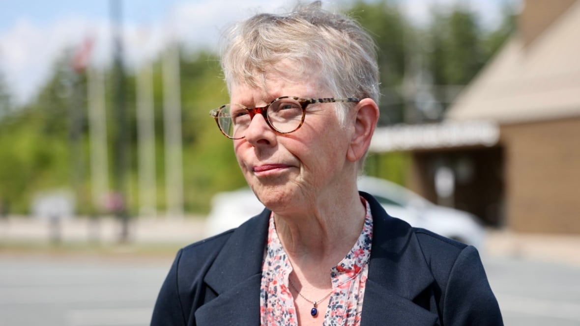 A achromatic pistillate with abbreviated grey hairsbreadth and glasses stands extracurricular a municipal gathering with emblem poles successful the background. She wears a flowered garment nether a navy blazer.