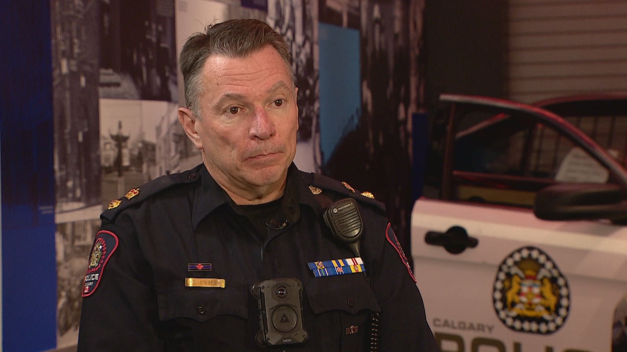 A man with short dark hair that's greying at the sides is wearing a police uniform sitting in front of a police cruiser with open doors.