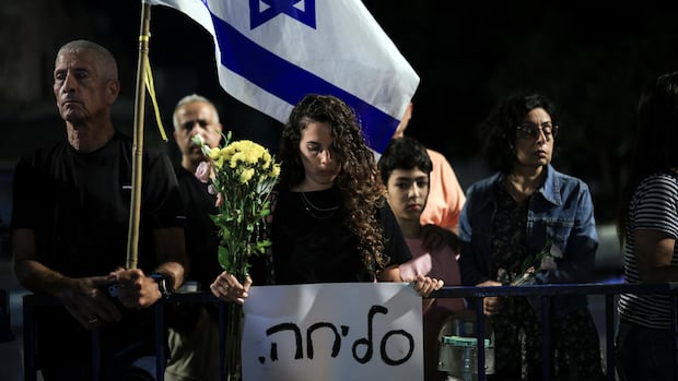 Solemn people stand near a barricade. One carries a sign, another the Israeli flag.