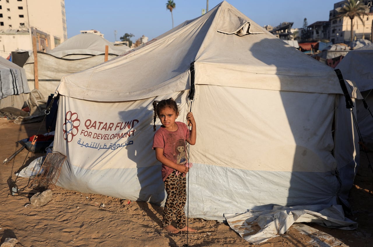 A young girl stands outside a canvas tent amid rubble.