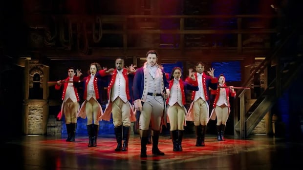 Wide shot of Hamilton cast on stage performing musical number. Actors are wearing 18th century costumes with colourful jackets.