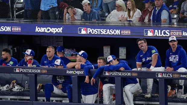 Baseball players wearing blue and white uniforms lean over the fence of a dugout inside a baseball stadium. 
