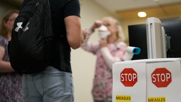A nurse demonstrates how to put on a mask at a measles screening point at Victoria Hospital in London, Ontario on July 9, 2025. Canada, which officially eradicated measles in 1998, has recorded more cases of the virus this year than any other Western country, according to World Health Organization figures updated this month. Different communities have been hit, but experts link the severity of the outbreak primarily to conservative Mennonite communities in the provinces of Ontario and Alberta long been opposed to vaccines.  People are stopped in a hospital with a sign that says "measles screening."