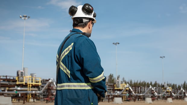 The Cenovus Christina Lake oil sands facility steam-assisted gravity drainage (SAGD) pad southeast of Fort McMurray, Alta., on Wednesday, April 24, 2024. A person in blue coveralls and hard hat stands in front of pipes.