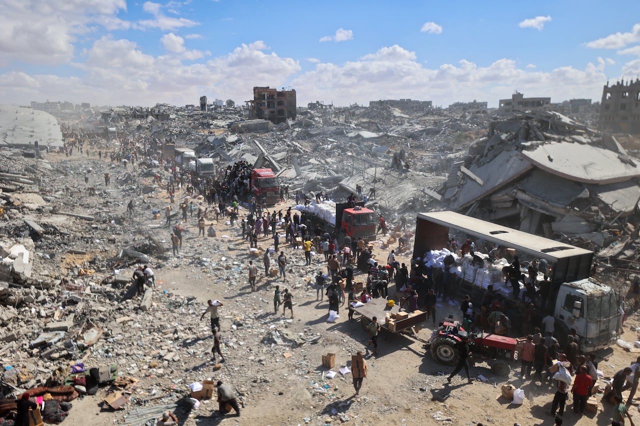 A destroyed CityScape, with houses and buildings reduced to rubble, can be seen as a line of real people with trucks bringing food and aid.