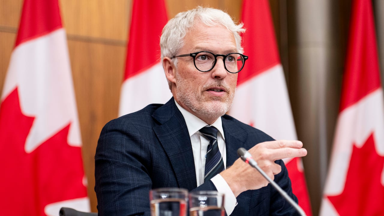Man sits at table with microphone and glasses of water in front of draped Canadian flags