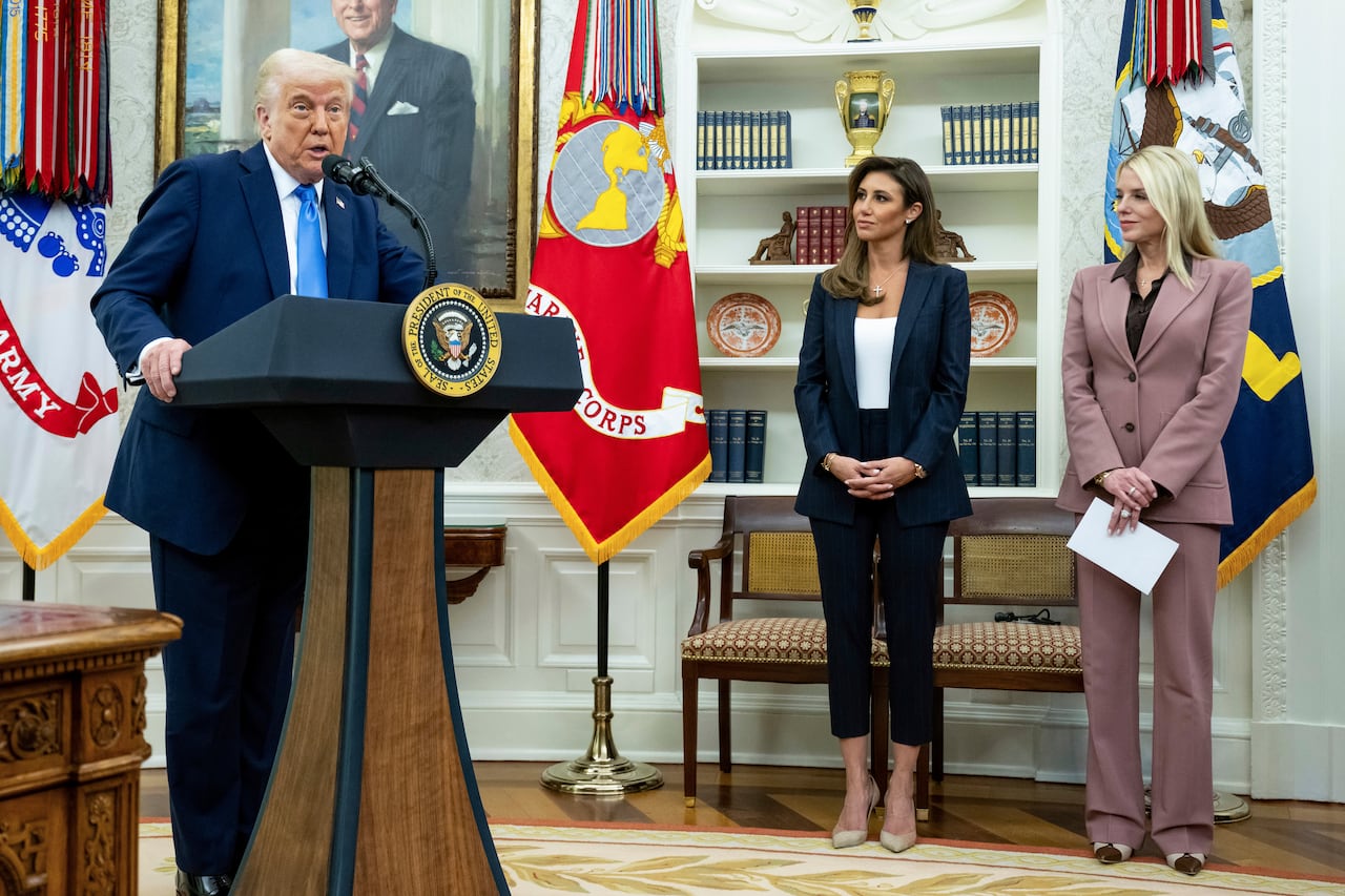 A neat old man in a suit and tie speaks from a podium in the office as two women in business suits, one blonde and one brunette, look.