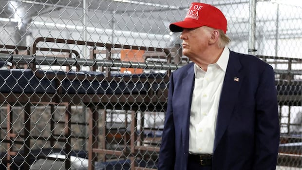 A man in a red cap stands beside bunk beds in a metal-fenced facility, looking contemplatively at his surroundings.