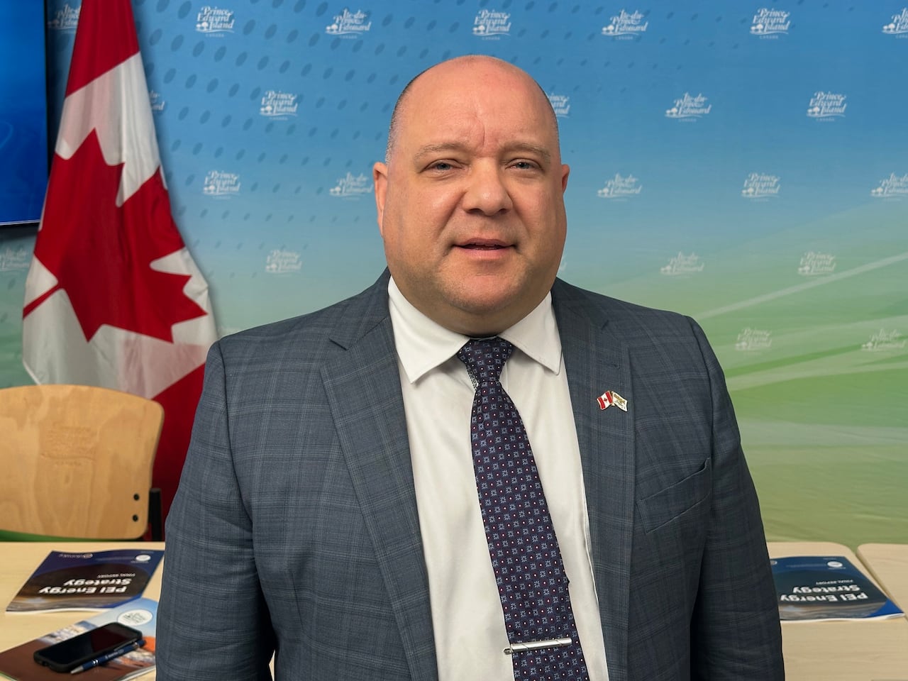 A man in a dark-coloured, plaid suit jacket and white shirt stands in front of a row of desks at an announcement.
