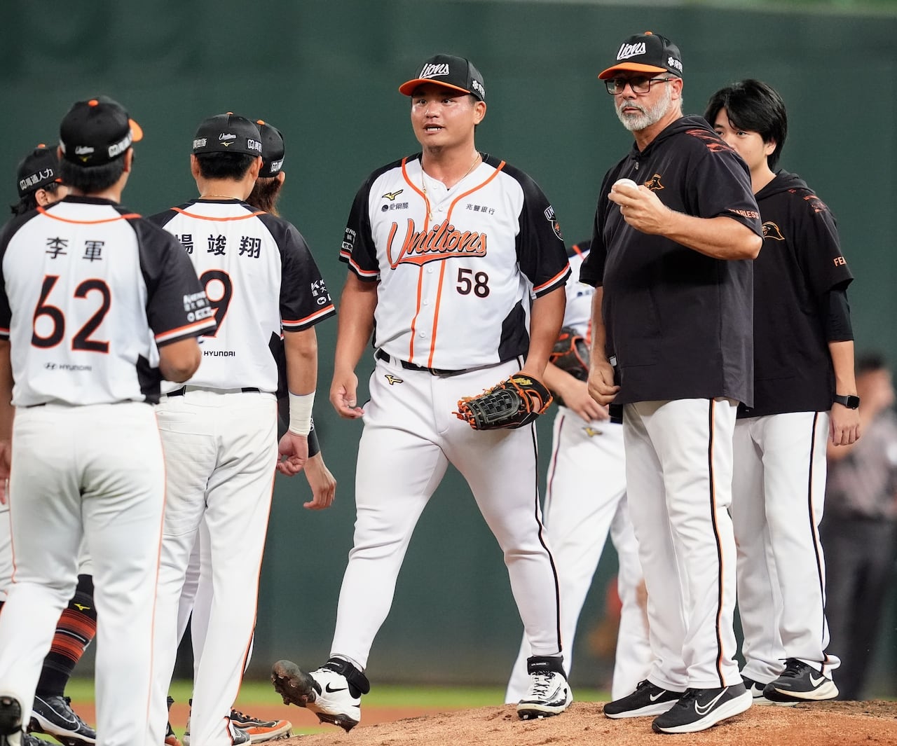 A pitching coach holds the baseball as several players gather on a mound.