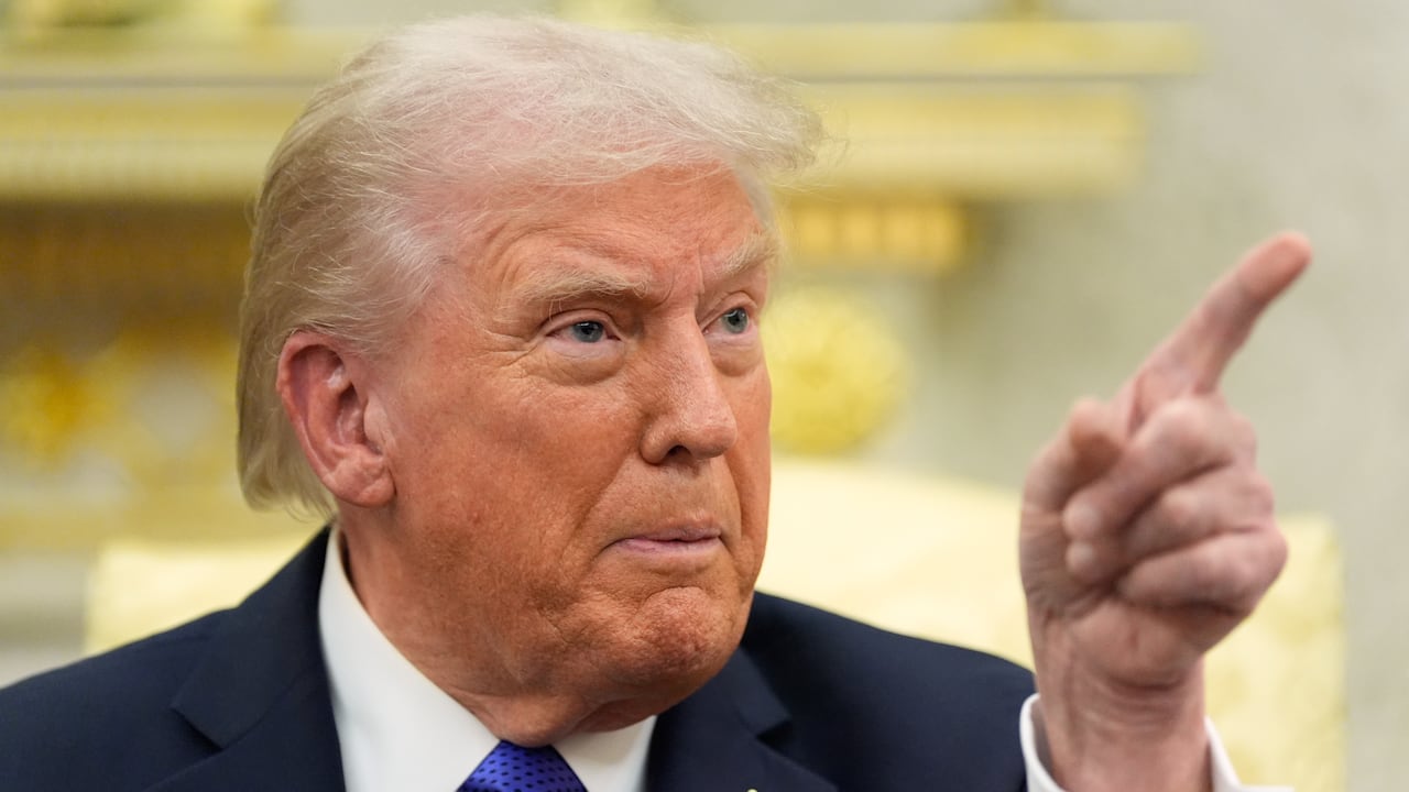 A man in a suit points in front of a golden backdrop. 