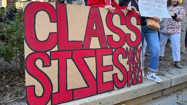 Several people stand outdoors beside a large size that says, 'Class sizes way too big.'