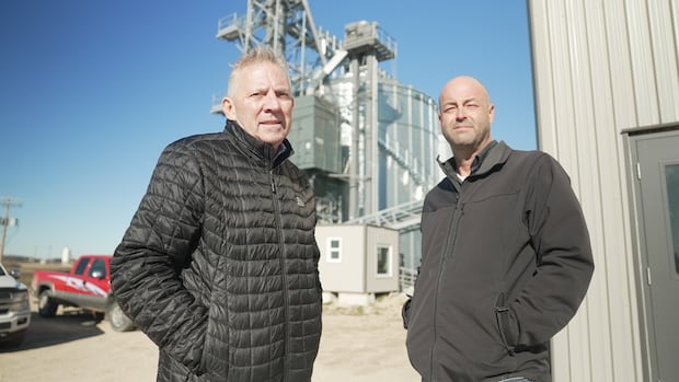 Two men in down puffy jackets standing in front of trucks and a grain elevator.