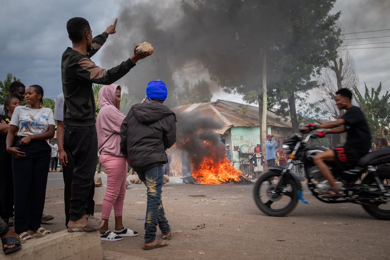 People in Tanzania stand around a burning fire, protesting. One man holds a stone in his raised hand
