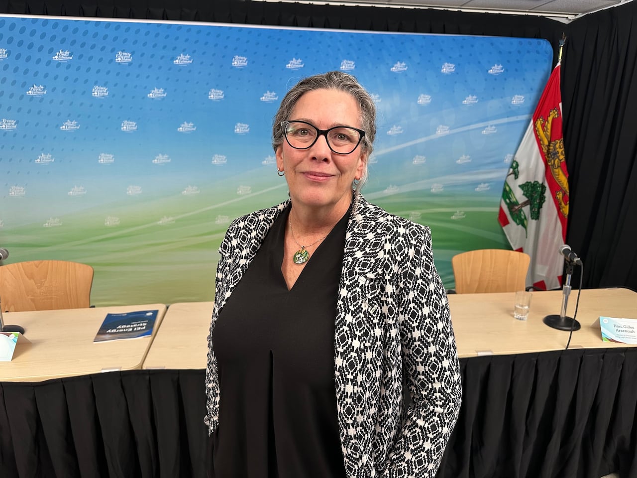 A woman in a black and white patterened blazer stands in front of a row of desks at an announcement.