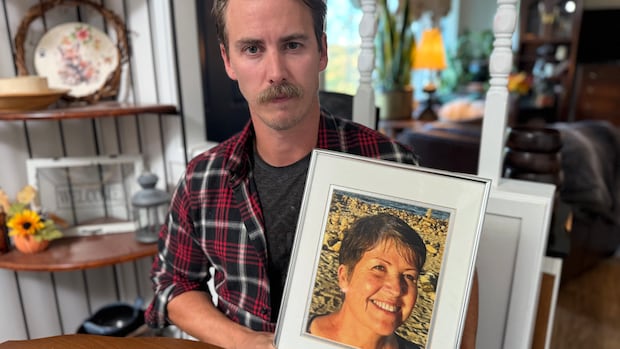 A man in his 30s with a moustache sits at his kitchen table, looking into the camera, holding a framed photo of a 69-year-old woman