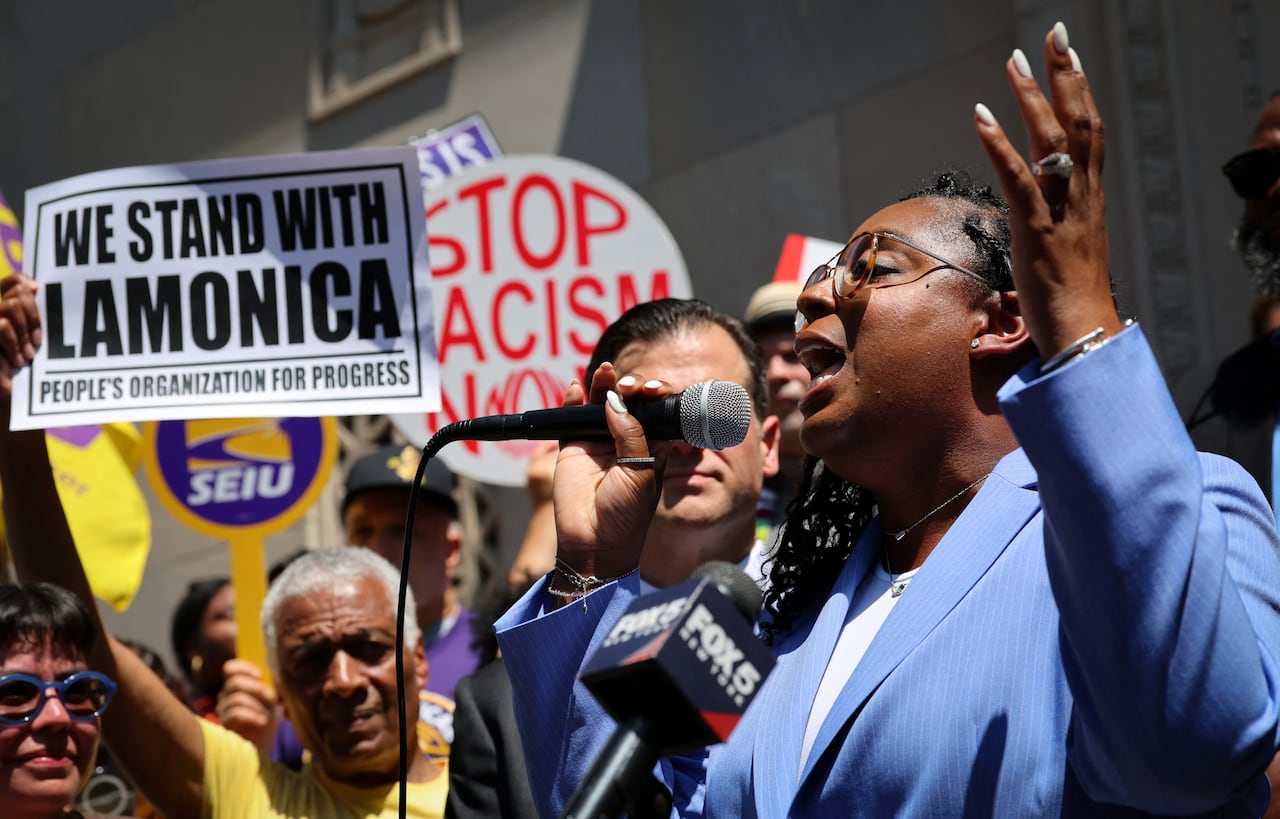 A dark-complected, bespectacled pistillate wearing a airy bluish blazer is shown successful illustration speaking into a microphone astatine an outdoor demonstration.