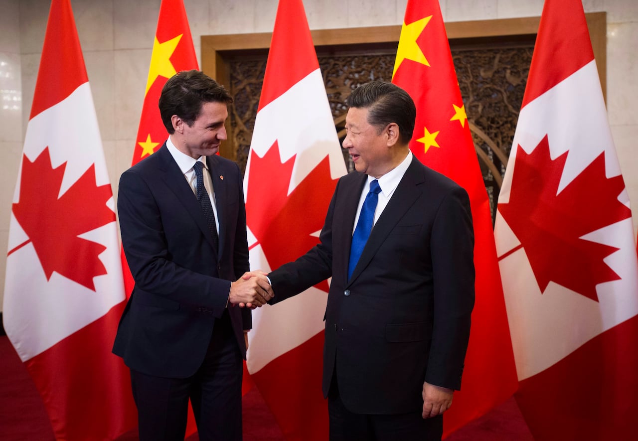 Two men in suits shake hands in front of a row of Canadian flags.