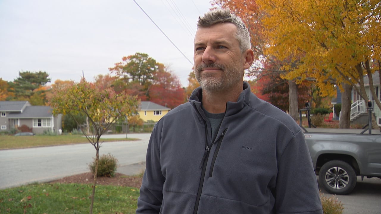 A man in a grey zip-up sweater stands with houses and trees visible behind him.